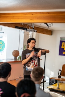 A man is standing in a room holding a microphone, seemingly speaking to an audience. He is wearing a graphic t-shirt and is positioned near a small table with water bottles and a basketball on it. The room has a modern feel, with wooden beams on the ceiling and various posters and signs on the walls. A few audience members are visible, focusing on him.