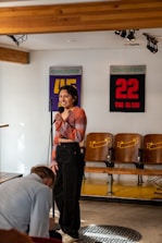 A person is standing and holding a microphone, seemingly performing or speaking in a casual setting. They are in a room with wooden beams on the ceiling and signs on the wall that read 'CONCOURSE 45' and '22 THE GLIDE'. Chairs with patterned backs are visible in the background, and there is adjustable lighting mounted on the ceiling.