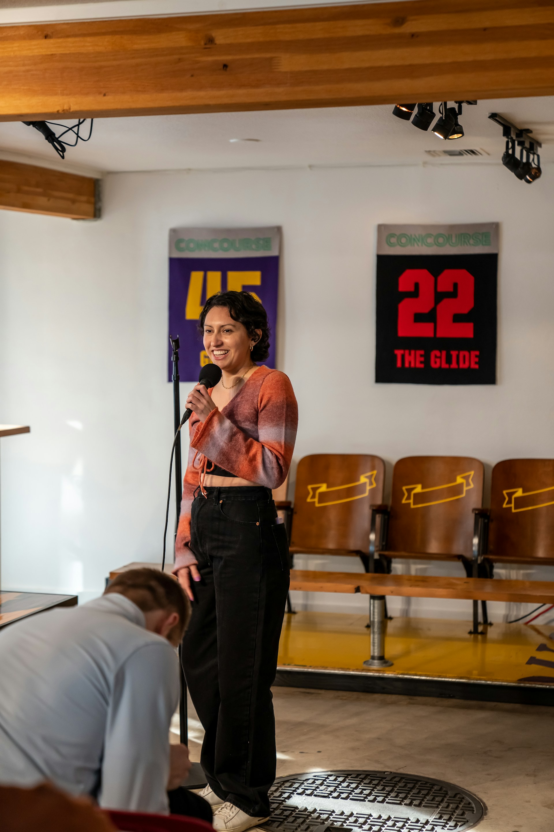 A person is standing and holding a microphone, seemingly performing or speaking in a casual setting. They are in a room with wooden beams on the ceiling and signs on the wall that read 'CONCOURSE 45' and '22 THE GLIDE'. Chairs with patterned backs are visible in the background, and there is adjustable lighting mounted on the ceiling.