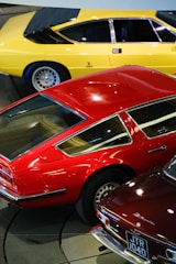 A collection of classic sports cars in vibrant colors, including a yellow car, a red car with retro styling, and another dark-colored car with a visible license plate. The cars are parked closely together in an indoor setting.