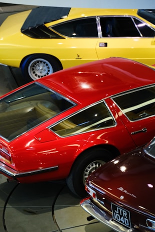 A collection of classic sports cars in vibrant colors, including a yellow car, a red car with retro styling, and another dark-colored car with a visible license plate. The cars are parked closely together in an indoor setting.