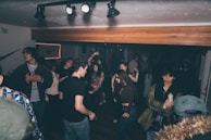 A lively indoor party scene with a group of people dancing and socializing under dim lighting. Some individuals are holding drinks, while others are engaged in conversations. The room has a casual atmosphere, with a neon sign visible in the background and spotlights on the ceiling.