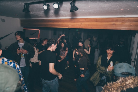 A lively indoor party scene with a group of people dancing and socializing under dim lighting. Some individuals are holding drinks, while others are engaged in conversations. The room has a casual atmosphere, with a neon sign visible in the background and spotlights on the ceiling.