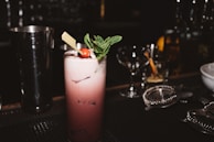 A dark cocktail bar setting features a tall, frothy cocktail topped with a sprig of mint and a cherry. The drink is served in a highball glass and garnished with a piece of chocolate. Various bar tools and glasses are in the background, creating a sophisticated atmosphere.