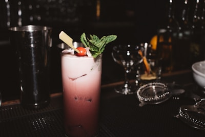A dark cocktail bar setting features a tall, frothy cocktail topped with a sprig of mint and a cherry. The drink is served in a highball glass and garnished with a piece of chocolate. Various bar tools and glasses are in the background, creating a sophisticated atmosphere.