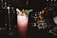 A dark cocktail bar setting features a tall, frothy cocktail topped with a sprig of mint and a cherry. The drink is served in a highball glass and garnished with a piece of chocolate. Various bar tools and glasses are in the background, creating a sophisticated atmosphere.
