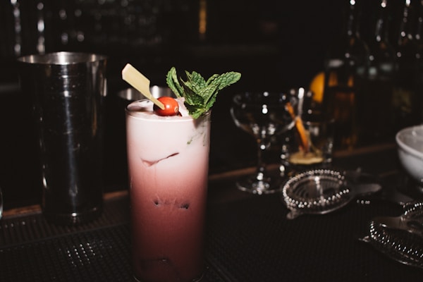A dark cocktail bar setting features a tall, frothy cocktail topped with a sprig of mint and a cherry. The drink is served in a highball glass and garnished with a piece of chocolate. Various bar tools and glasses are in the background, creating a sophisticated atmosphere.