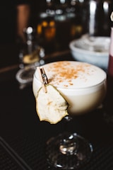A cocktail glass filled with a creamy beverage, topped with foam and sprinkled with spices. A dried fruit slice is clipped to the glass with a mini clothespin, adding an artistic touch. The background is softly blurred, with hints of a bar setting.