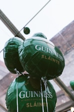 Green balloons with 'Guinness' and 'Sláinte' printed on them are clustered together. They are floating indoors and surrounded by a glass ceiling and a building structure in the background.