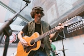 A musician wearing sunglasses and a hat is playing a guitar at an outdoor event. There are microphones and banners in the background, and another individual is partially visible behind him.