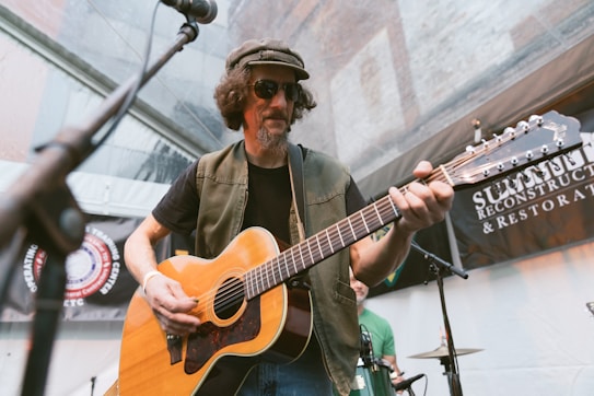 A musician wearing sunglasses and a hat is playing a guitar at an outdoor event. There are microphones and banners in the background, and another individual is partially visible behind him.