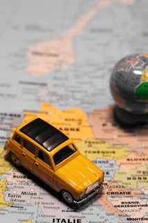 A small, vintage yellow toy car is placed on a map of Europe, highlighting cities in countries like Italy, France, and Switzerland. A small globe is partially visible on the right, emphasizing travel and exploration themes.
