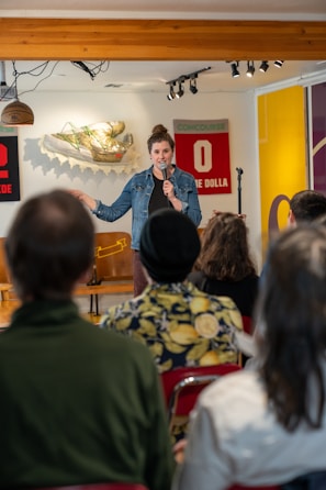 A woman holding a microphone stands on stage in front of an audience. She appears to be performing or speaking. There are decorative wall elements behind her, including a shoe sculpture and sports-themed items. The setting seems casual and intimate.