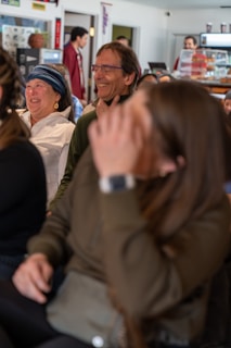 A group of people sitting together in a casual setting, likely a café or informal meeting space. The focus is on two individuals, a woman wearing a blue headband and a man with glasses, both appearing to be sharing a joyful moment. Other people are visible in the background near a counter.