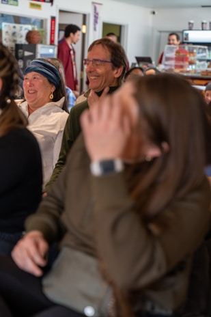 A group of people sitting together in a casual setting, likely a café or informal meeting space. The focus is on two individuals, a woman wearing a blue headband and a man with glasses, both appearing to be sharing a joyful moment. Other people are visible in the background near a counter.