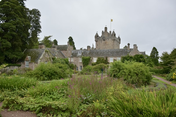 A historic stone manor house surrounded by lush greenery and an expansive, well-maintained garden. The structure features a tower with a flagpole and is set against a backdrop of mature trees. The garden is filled with various types of plants, shrubs, and flowers, adding to the scenic beauty of the estate.