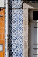 Detailed blue and white patterned tiles are adorning a wall, featuring intricate designs next to a section of exposed beige stone. A vertical metal pipe runs along the left side of the wall. Adjacent sections of the wall feature orange tiles, creating a contrast with the patterned tiles.
