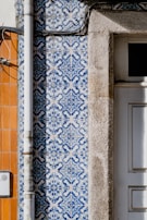 Detailed blue and white patterned tiles are adorning a wall, featuring intricate designs next to a section of exposed beige stone. A vertical metal pipe runs along the left side of the wall. Adjacent sections of the wall feature orange tiles, creating a contrast with the patterned tiles.
