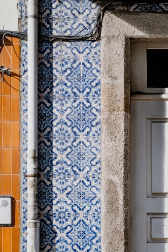Detailed blue and white patterned tiles are adorning a wall, featuring intricate designs next to a section of exposed beige stone. A vertical metal pipe runs along the left side of the wall. Adjacent sections of the wall feature orange tiles, creating a contrast with the patterned tiles.