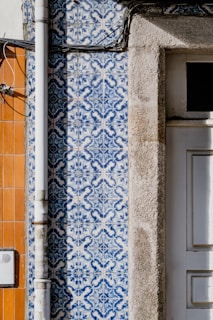 Detailed blue and white patterned tiles are adorning a wall, featuring intricate designs next to a section of exposed beige stone. A vertical metal pipe runs along the left side of the wall. Adjacent sections of the wall feature orange tiles, creating a contrast with the patterned tiles.