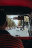 A view from inside a vehicle, looking out onto a street. Several rickshaws are visible on the road, with one prominently in the center. There are buildings and trees lining the street, and a person can be seen partially from behind.