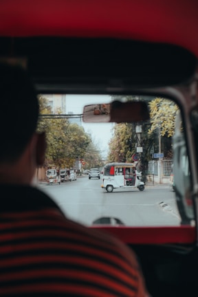 A view from inside a vehicle, looking out onto a street. Several rickshaws are visible on the road, with one prominently in the center. There are buildings and trees lining the street, and a person can be seen partially from behind.