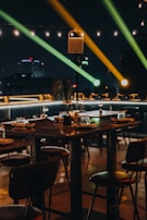 A dimly lit outdoor dining area with wooden tables and chairs is set for guests. The ambiance is enhanced by overhead string lights and colorful spotlights creating an inviting and warm atmosphere. The backdrop includes a cityscape silhouetted against the night sky.