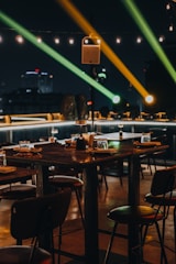 A dimly lit outdoor dining area with wooden tables and chairs is set for guests. The ambiance is enhanced by overhead string lights and colorful spotlights creating an inviting and warm atmosphere. The backdrop includes a cityscape silhouetted against the night sky.