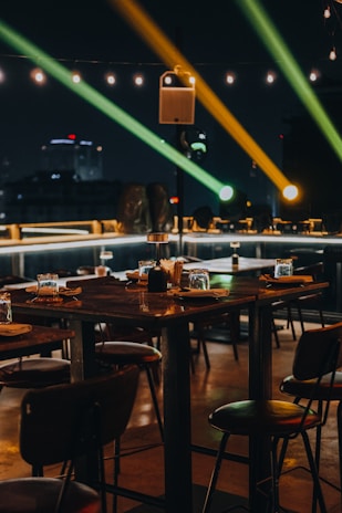 A dimly lit outdoor dining area with wooden tables and chairs is set for guests. The ambiance is enhanced by overhead string lights and colorful spotlights creating an inviting and warm atmosphere. The backdrop includes a cityscape silhouetted against the night sky.