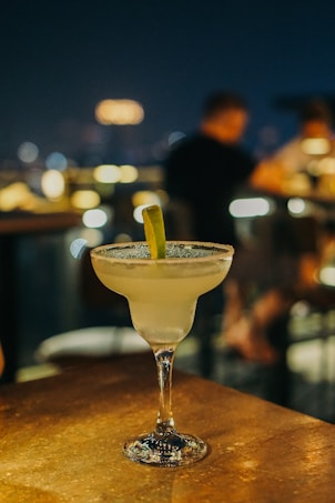 A margarita cocktail glass with a salted rim and a lime wedge garnish sits on a wooden table in a dimly lit bar setting. The background features blurred figures of people and bokeh lights, suggesting a lively evening atmosphere.