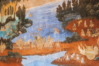 A traditional mural painting depicting a historical or mythological scene with detailed characters and landscapes. The setting includes a flowing body of water, lush vegetation, and numerous figures adorned in elaborate attire engaged in various activities. The colors are rich and earthy, suggesting a scene from cultural folklore.