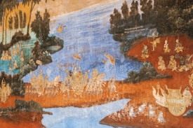 A traditional mural painting depicting a historical or mythological scene with detailed characters and landscapes. The setting includes a flowing body of water, lush vegetation, and numerous figures adorned in elaborate attire engaged in various activities. The colors are rich and earthy, suggesting a scene from cultural folklore.