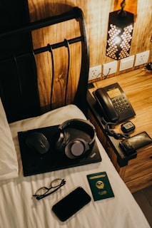 A cozy bedroom scene featuring a neatly arranged wooden nightstand with a stylish lamp, a telephone, and electronic devices. On the bed, a laptop is accompanied by a pair of headphones, a mouse, a pair of glasses, and a smartphone next to an international passport.