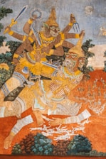 A detailed fresco depicts a mythical battle scene with two vividly dressed figures, both wearing traditional Southeast Asian attire. The figure on top has multiple arms, holding various weapons including a sword and a discus. The background features mystical landscapes with trees and a sky with a moon.