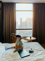 A young child wearing headphones sits on a bed in a room with large windows showing a cityscape. The child is dressed casually in a gray shirt and colorful socks. Next to the child on the bed are a laptop and a vintage camera. There is a small round wooden table by the window with a chair and a cup on top, and the curtains are open letting in bright natural light.
