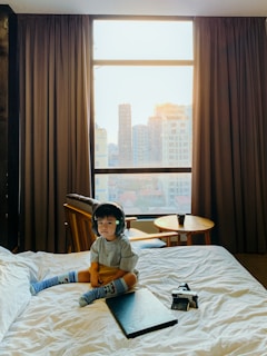 A young child wearing headphones sits on a bed in a room with large windows showing a cityscape. The child is dressed casually in a gray shirt and colorful socks. Next to the child on the bed are a laptop and a vintage camera. There is a small round wooden table by the window with a chair and a cup on top, and the curtains are open letting in bright natural light.