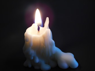 A single, partially melted white candle with a visible flame burning steadily against a dark background. The wax appears to have dripped down, forming intricate patterns along its sides.