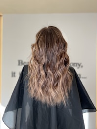 Long, wavy brown hair with soft curls cascading down the back, combined with a black cape typically found in hair salons. The background is slightly blurred with indistinct text visible.