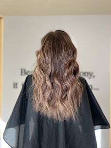 Long, wavy brown hair with soft curls cascading down the back, combined with a black cape typically found in hair salons. The background is slightly blurred with indistinct text visible.