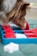 A dog is closely examining or playing with a puzzle toy, which consists of red and white pieces on a blue base. The setting seems to be an indoor space with a green cushioned surface beneath the toy.