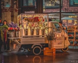 A small, charming flower truck with metal buckets full of colorful flowers is parked indoors. The truck is adorned with a decorative sign, and a woman stands nearby examining the vibrant floral arrangement. The setting suggests a market or indoor event space with exposed brick walls and large windows letting in natural light.