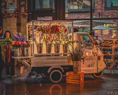 A small, charming flower truck with metal buckets full of colorful flowers is parked indoors. The truck is adorned with a decorative sign, and a woman stands nearby examining the vibrant floral arrangement. The setting suggests a market or indoor event space with exposed brick walls and large windows letting in natural light.