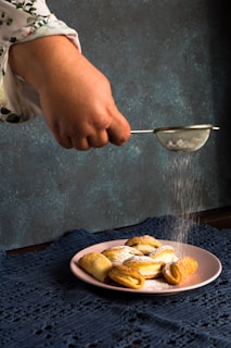 A hand is sprinkling powdered sugar over a plate of pastries. The setup is on a textured dark blue surface with a moody backdrop. The pastries, arranged on a pink plate, look golden brown and freshly baked. The action of dusting enhances the visual appeal, creating a sense of homemade cooking.