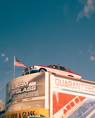 A vintage car is mounted on top of a building with a large advertisement for an alarm and glass company. An American flag is flying beside the car. The building displays various promotional text and logos, with one side featuring bold red and blue graphics. The sky is clear with a deep blue color.