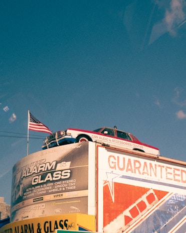 A vintage car is mounted on top of a building with a large advertisement for an alarm and glass company. An American flag is flying beside the car. The building displays various promotional text and logos, with one side featuring bold red and blue graphics. The sky is clear with a deep blue color.