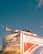 A vintage car is mounted on top of a building with a large advertisement for an alarm and glass company. An American flag is flying beside the car. The building displays various promotional text and logos, with one side featuring bold red and blue graphics. The sky is clear with a deep blue color.