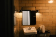 A dimly lit bathroom scene with beige tiled walls. A rectangular mirror is mounted above a white ceramic sink. There are two wall-mounted lights above the mirror, casting a warm glow. A metal stand holds a roll of paper towels and a glass.