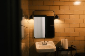 A dimly lit bathroom scene with beige tiled walls. A rectangular mirror is mounted above a white ceramic sink. There are two wall-mounted lights above the mirror, casting a warm glow. A metal stand holds a roll of paper towels and a glass.