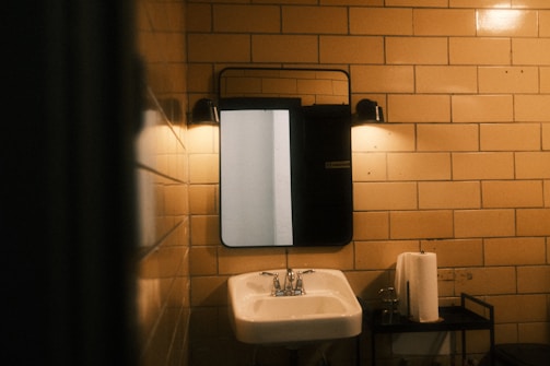A dimly lit bathroom scene with beige tiled walls. A rectangular mirror is mounted above a white ceramic sink. There are two wall-mounted lights above the mirror, casting a warm glow. A metal stand holds a roll of paper towels and a glass.