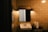 A dimly lit bathroom scene with beige tiled walls. A rectangular mirror is mounted above a white ceramic sink. There are two wall-mounted lights above the mirror, casting a warm glow. A metal stand holds a roll of paper towels and a glass.